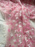 a close-up view of a fabric with a pink and white floral pattern. The fabric appears to be made of sheer material, with a delicate and intricate design. The pattern consists of numerous small white flowers interspersed with pink flowers, creating a visually appealing and delicate appearance. The fabric is spread out, revealing its texture and the way the flowers are arranged on it.