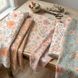 a collection of fabric swatches with floral patterns, including pink, white, and light blue colors, arranged on a wooden table. There are also sewing supplies, such as scissors and a bowl, visible in the scene.