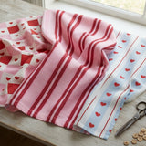 a wooden table with three colorful fabric pieces, including a pink and white striped fabric, a blue and white striped fabric, and a red and white striped fabric. There are also some scissors and buttons on the table.