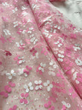 a close-up view of a fabric with a delicate floral pattern in shades of pink and white. The fabric appears to be sheer, with small white flowers or butterflies scattered across the surface, creating a soft and delicate appearance.