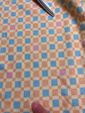 a close-up view of a fabric with a checkered pattern in shades of blue, orange, and pink. A pair of scissors is visible on the left side of the image.